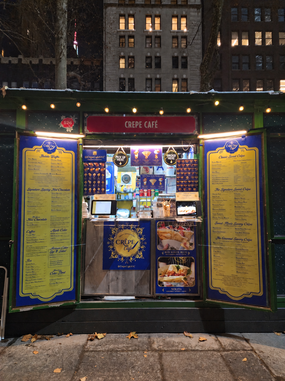 The front of Crêpe Café kiosk S26 at night, with open blue-and-gold menu doors and warm string lights